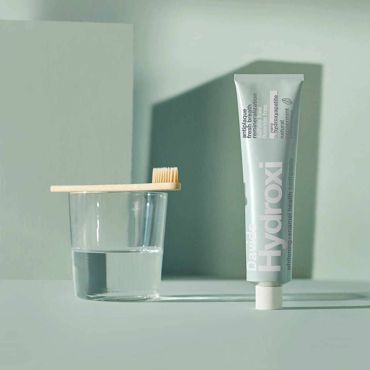 Tube of Davids Hydroxi whitening enamel health nano hydroxyapatite toothpaste with bamboo toothbrush and water glass on green background