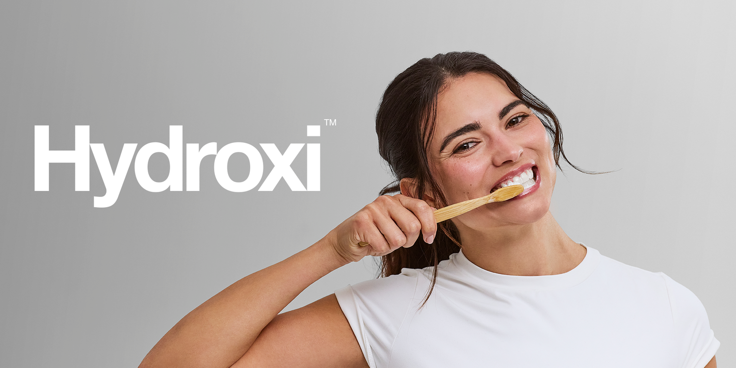 Woman brushing teeth with bamboo toothbrush next to Hydroxi logo on gray background