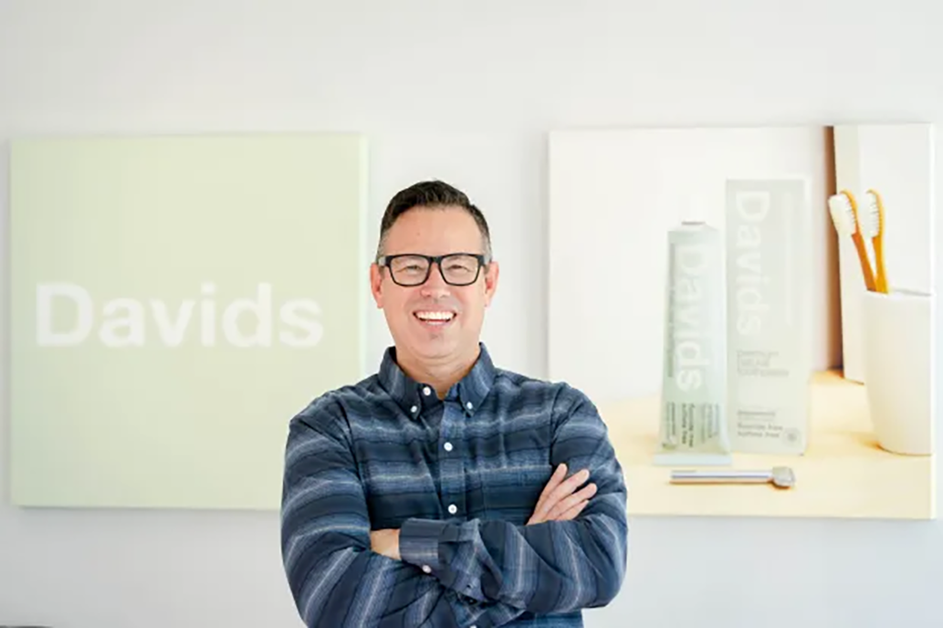 Founder Eric Buss smiling with arms crossed, standing in front of Davids toothpaste posters