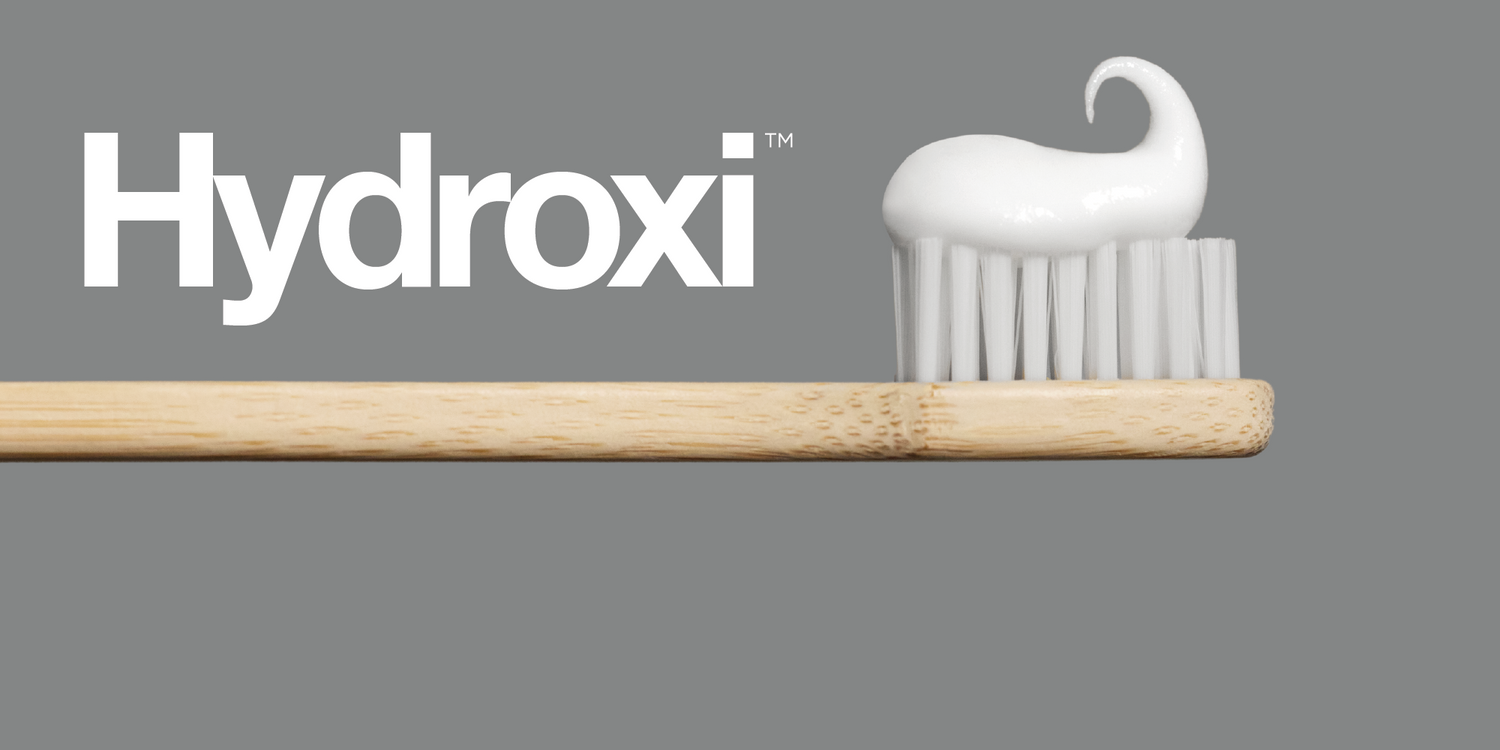 Toothpaste swirl on white-bristle bamboo toothbrush with "Hydroxi" text on gray background