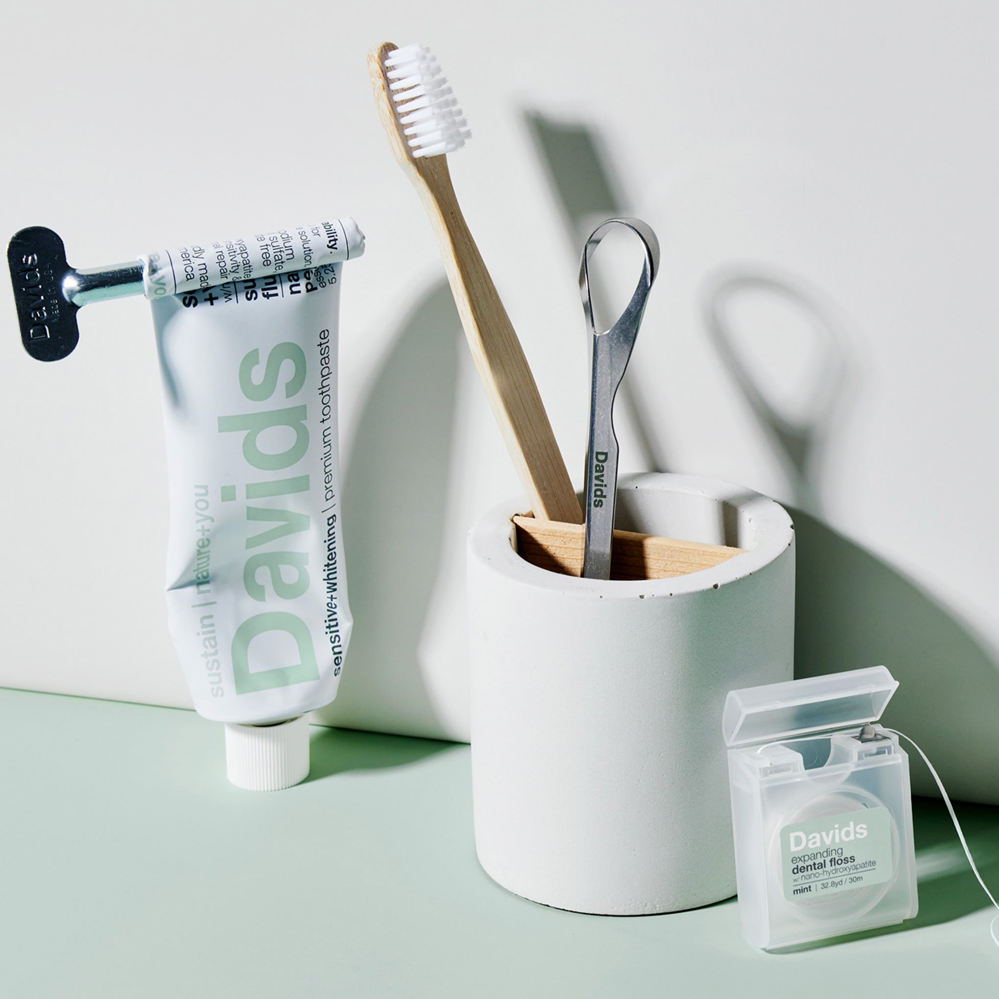 Davids toothpaste, bamboo toothbrush, tongue scraper in holder, and mint dental floss on pale surface