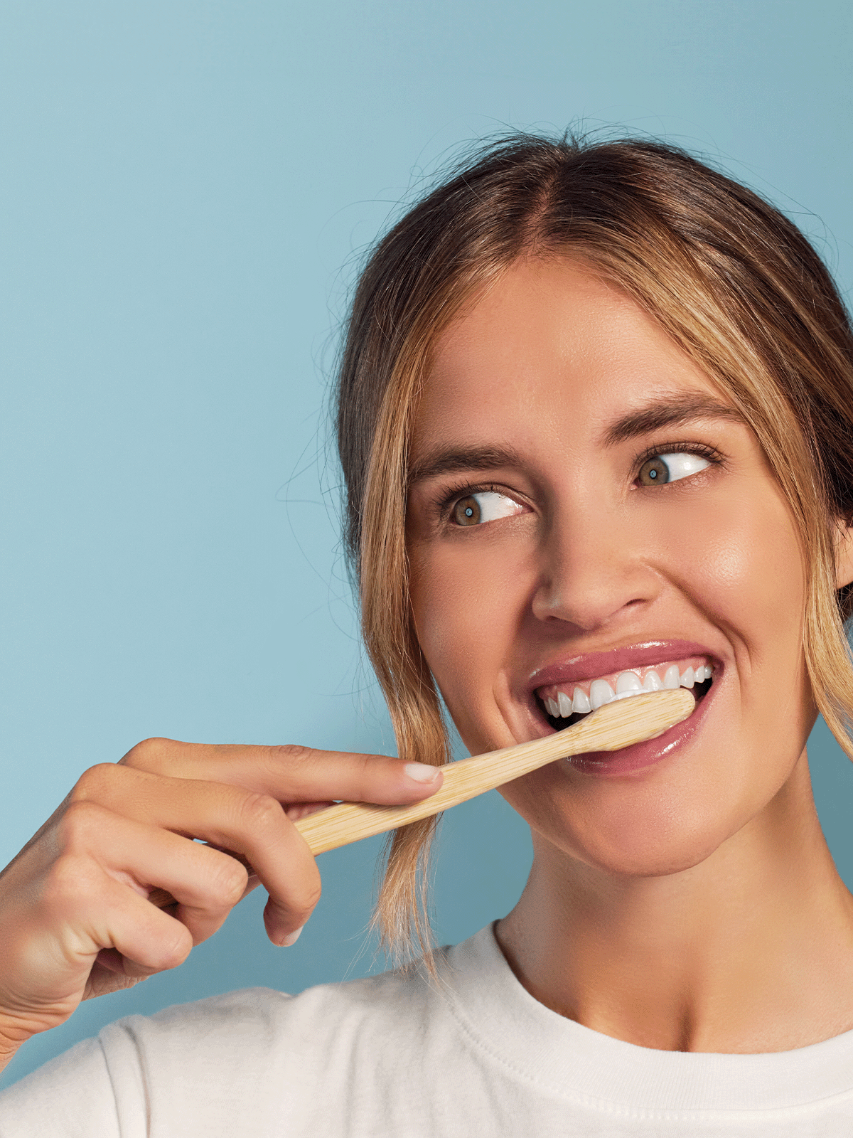 Woman brushing teeth with a bamboo toothbrush, smiling against a light blue background