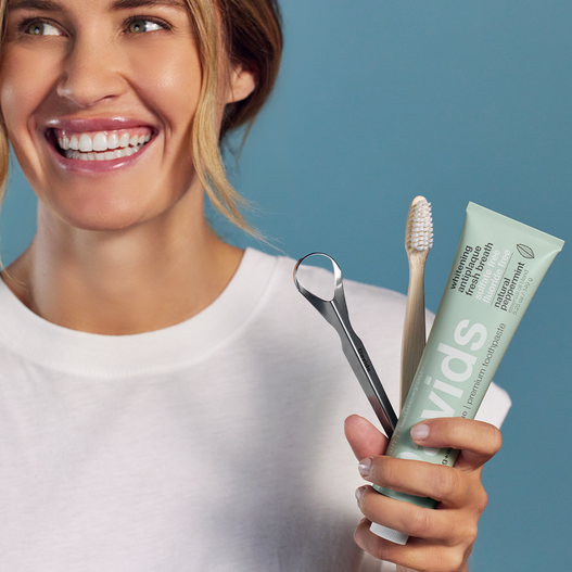 Smiling woman holding Davids toothpaste, a wooden toothbrush, and a metal tongue scraper