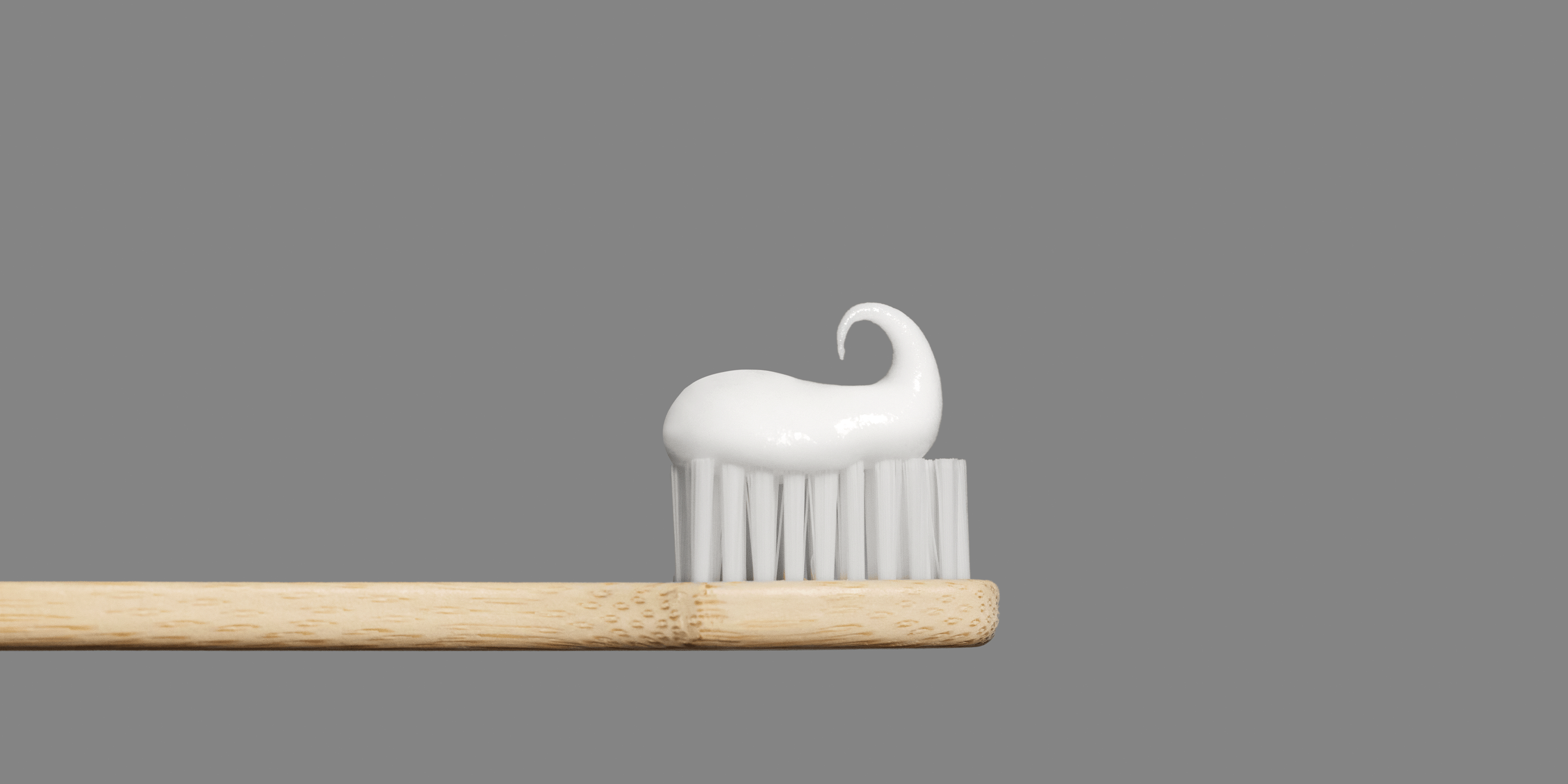 Wooden toothbrush with a swirl of white toothpaste on the bristles against a gray background